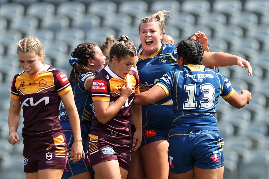An Eels NRLW player stands calling out in celebration as her teammates come in past dejected Broncos players.