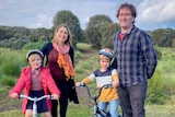 A mother and father stand beside their two children. The kids are on bikes in a park.