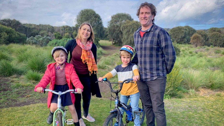 A mother and father stand beside their two children. The kids are on bikes in a park.