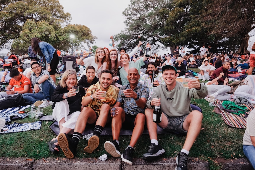 A group of people sit on a grassy hill amid large crowds, all holding drinks and smiling. 