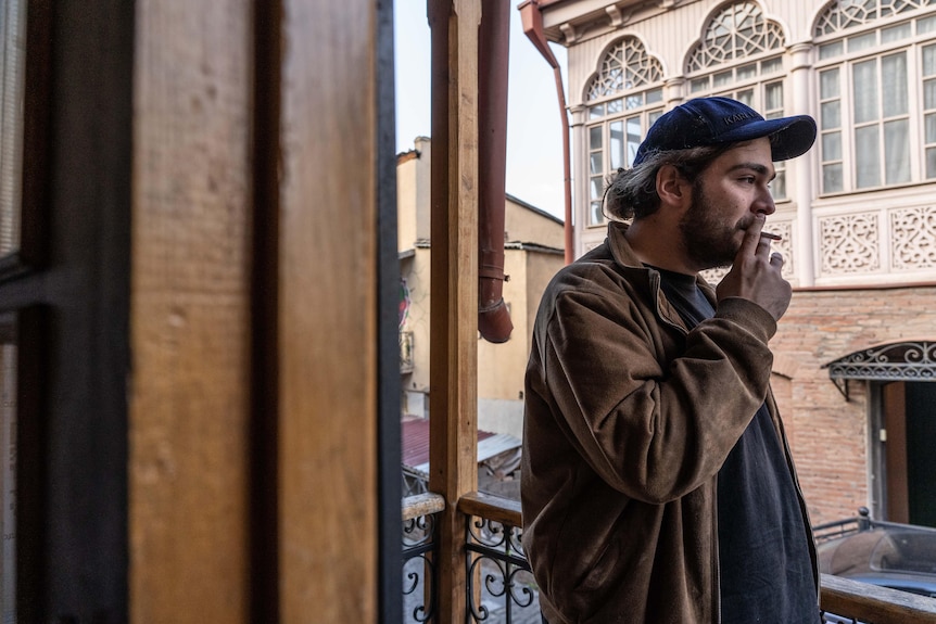 A man in a blue cap is smoking on a balcony.  The buildings behind him are ornate and beautiful