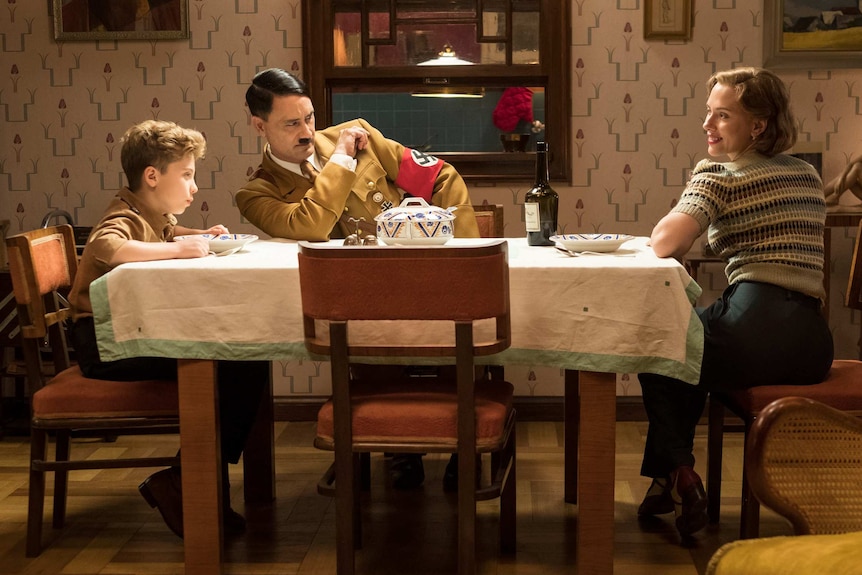A boy, Hitler character, and woman sit at the dinner table in a carefully arranged room full of square edges
