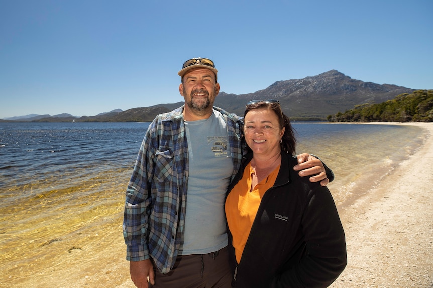 A man and a woman stand together on a beach with mountains in the background