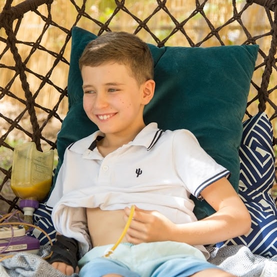 A 10-year-old boy sits on outdoor swing chair, with a feeding device attached to his stomach.