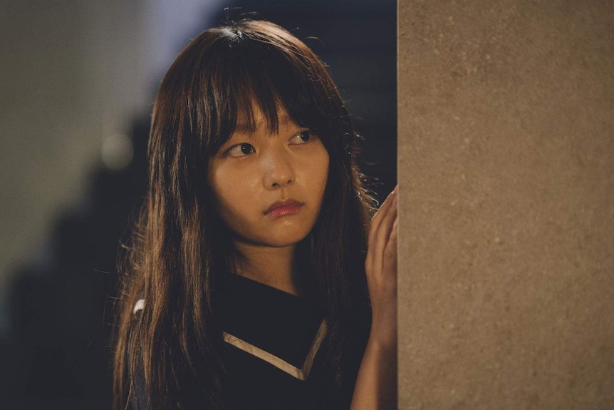 A young woman with long hair and a blunt fringe peeks out from behind a wall.