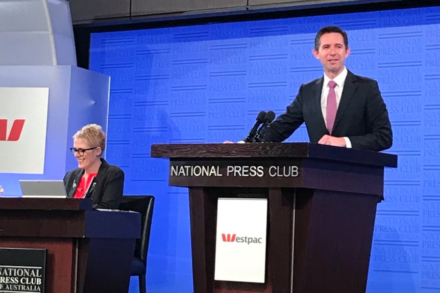 Simon Birmingham standing behind a brown lectern wearing a black suit