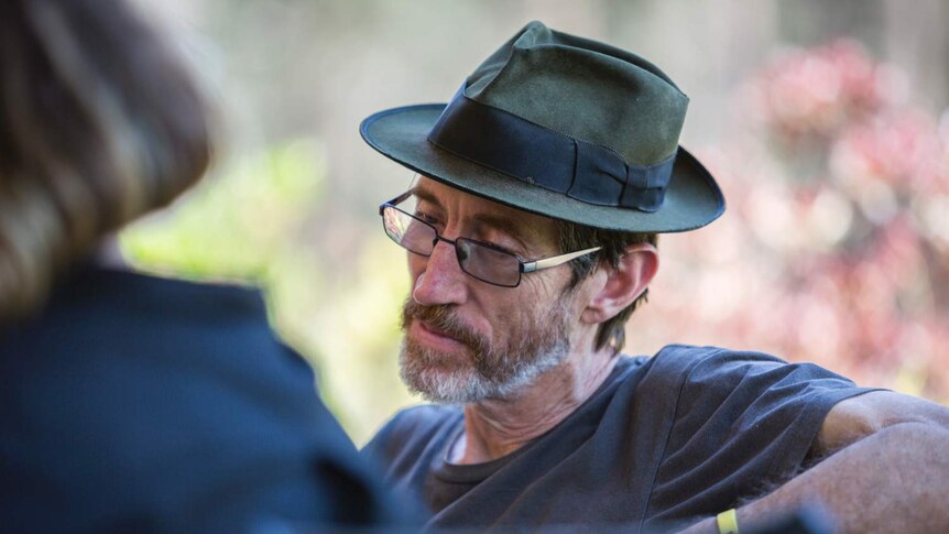John Ellis, wearing glasses and a hat, sits on an outdoor bench.