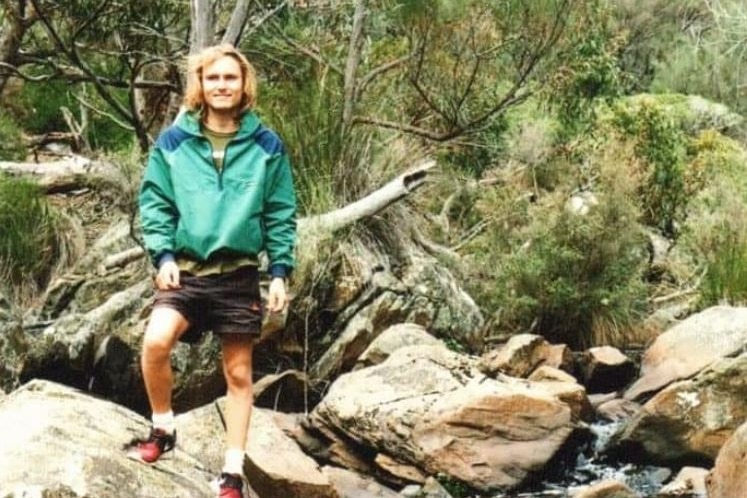 A young man with a green jacket and shoulder-length blonde hair stands on rocks