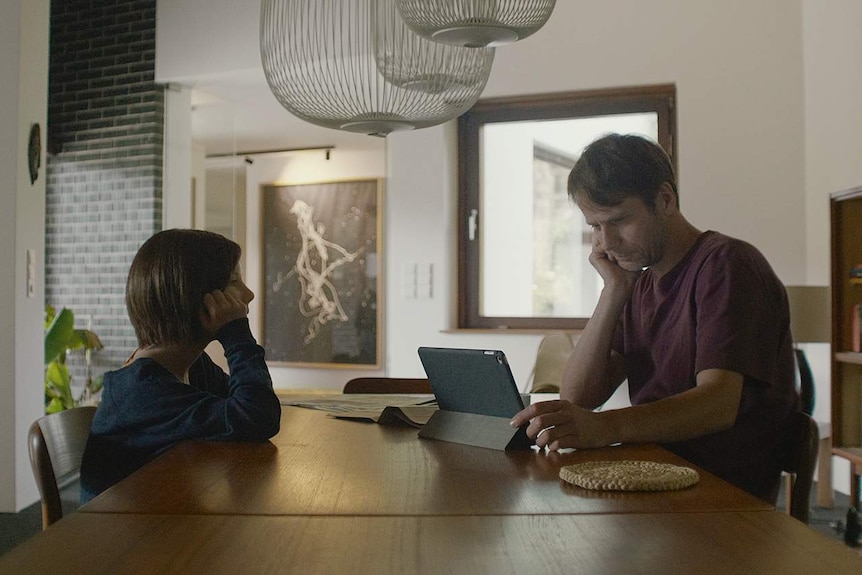 A man and a young AI girl sit at a table in a house in the film The Trouble with Being Born