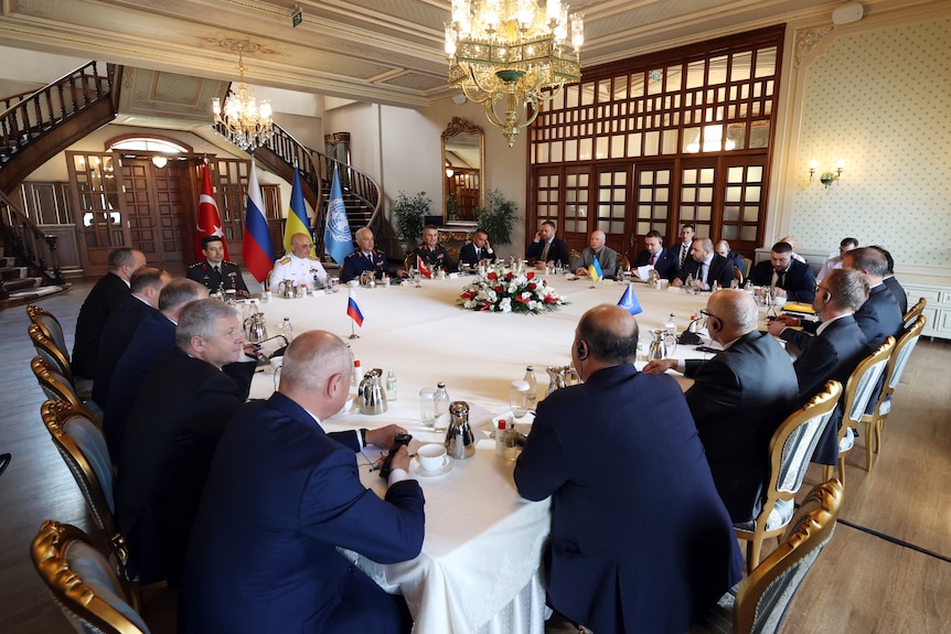 Mężczyźni w formalnych ubraniach siedzą wokół dużego kwadratowego stołu z flagami Turcji, Rosji, Ukrainy i ONZ w tle.