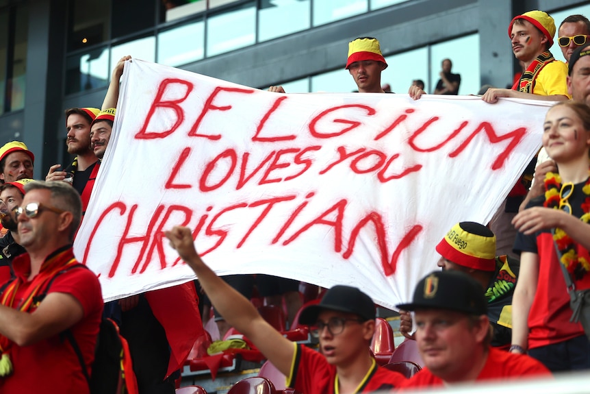 A white banner saying "Belgium loves you Christian" in red letters is held up by supporters