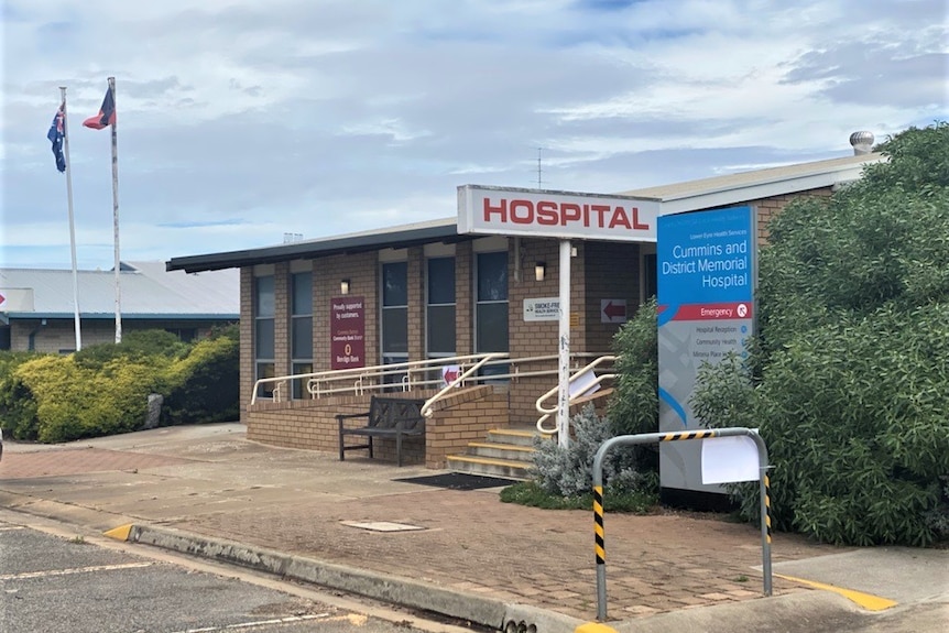 The front of a brown brick building with a HOSPITAL sign in red & a blue sign with the name next to a bush. 2 flags in backroud