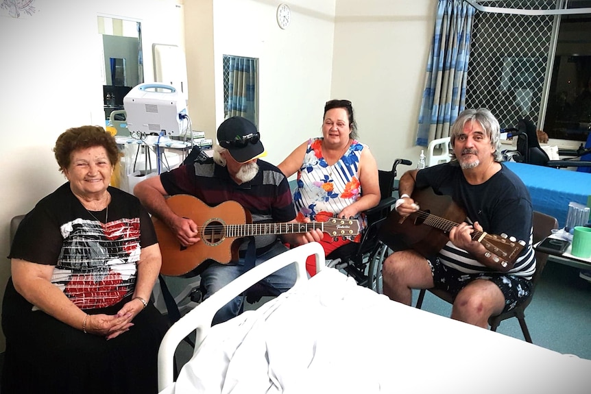A group of two men and two women sit around a hospital bed playing the guitar