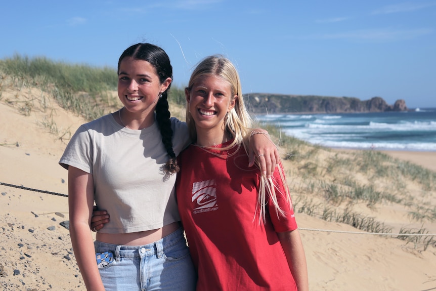 Two girls stand in front of beach with cliffs in the background, arms around one another.