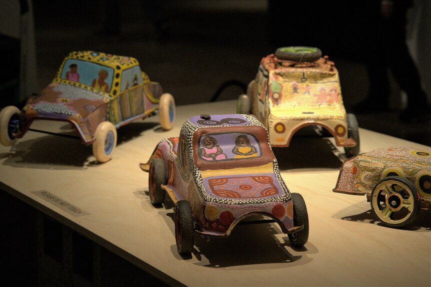 Sculptures of cars with Indigenous dot paintings on them sit under dim down lights in an art gallery exhibit