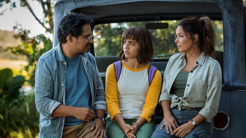A young girl with backpack looks concerned and sits in between her parents, a man and woman, on the back of motor vehicle.