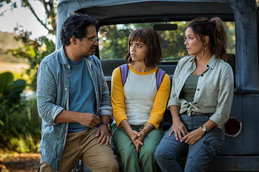 A young girl with backpack looks concerned and sits in between her parents, a man and woman, on the back of motor vehicle.