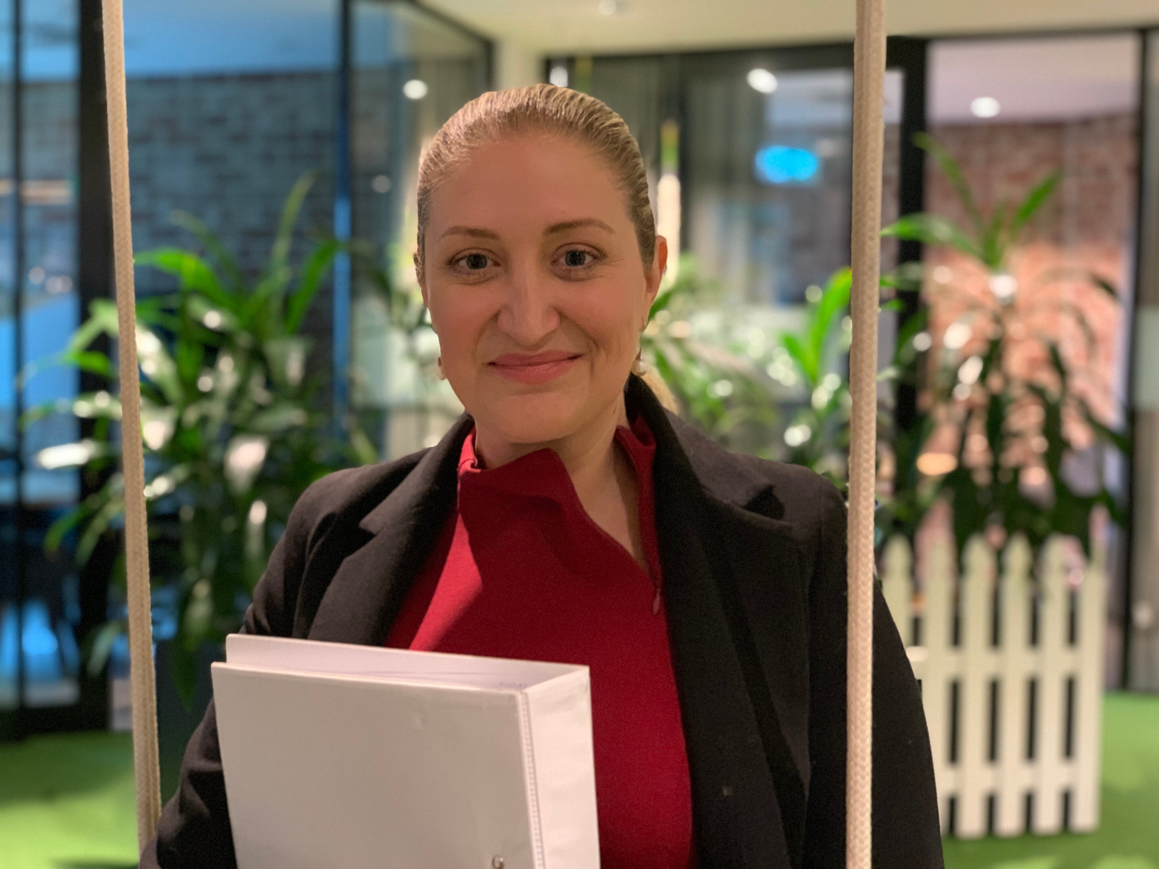 A woman in a red shirt and black jacket smiles, holding a binder in her arms.