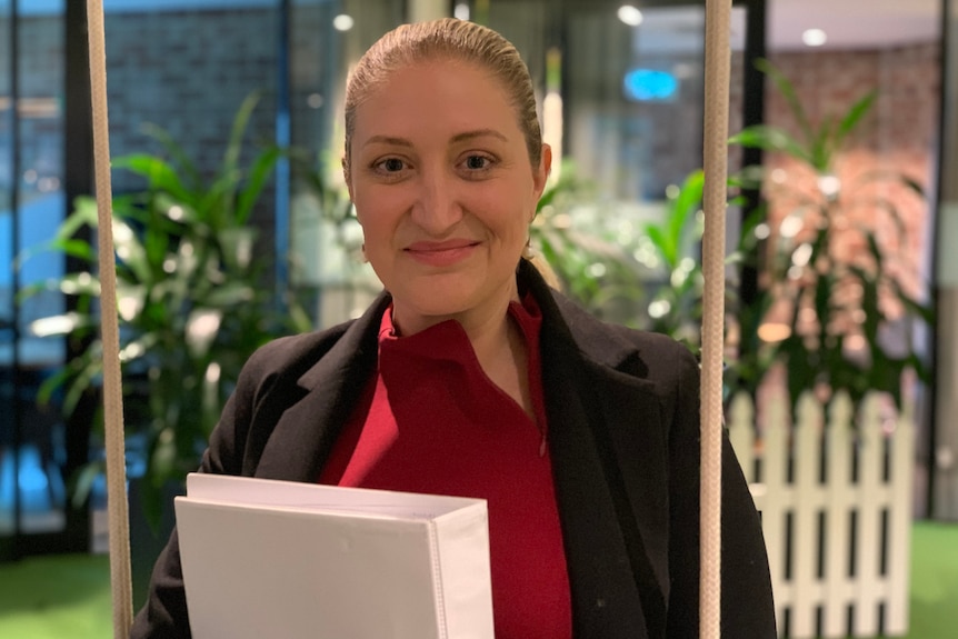 A woman in a red shirt and black jacket smiles, holding a binder in her arms.