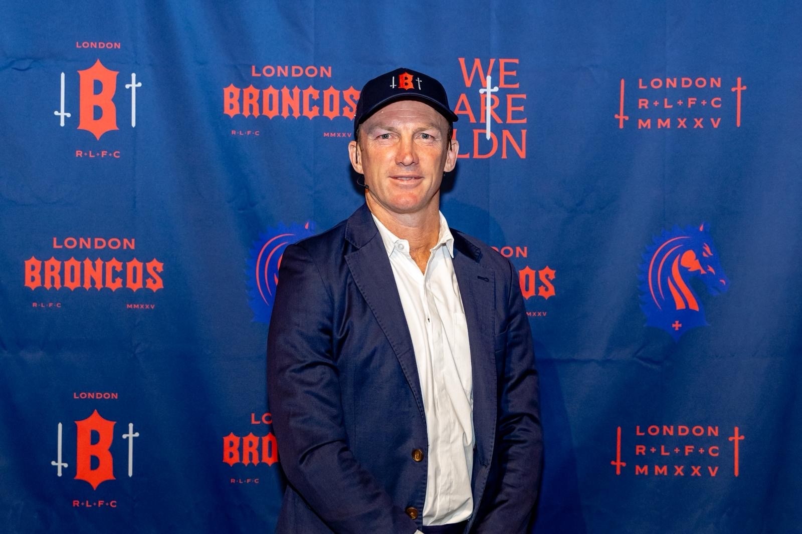 A man in a dark suit wears a cap as he stands in front of a backdrop with "London Broncos" branding.