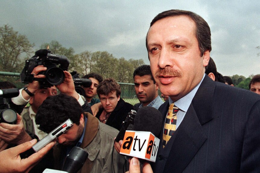 A 1990s photo of a Turkish man in a suit, surrounded by reporters.