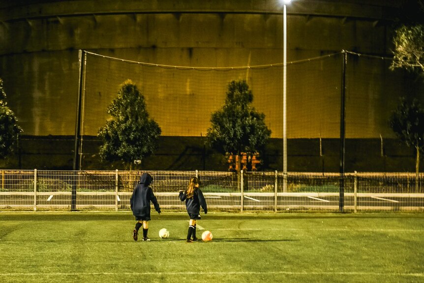 Two children walk along a soccer pitch dribbling balls, with an oil tank behind them