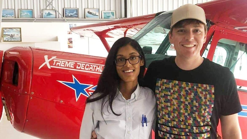 A couple smile at the camera with a plane behind them.