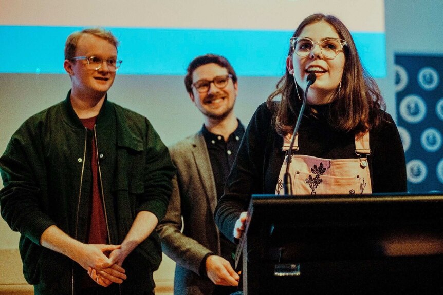 Grace Bruxner speaking into a microphone and two men in the background standing on a stage