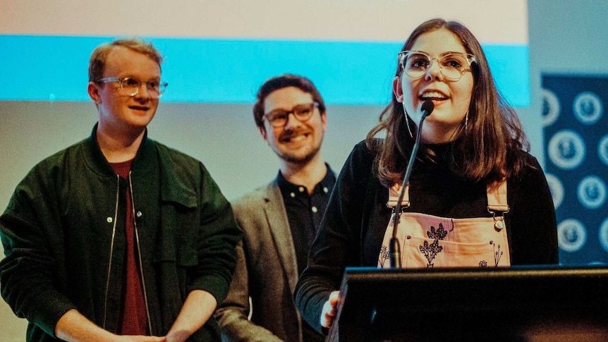 Grace Bruxner speaking into a microphone and two men in the background standing on a stage