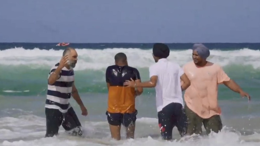 Four men, two of whom are wearing turbans, stand knee-deep in the ocean as the surf rolls in.