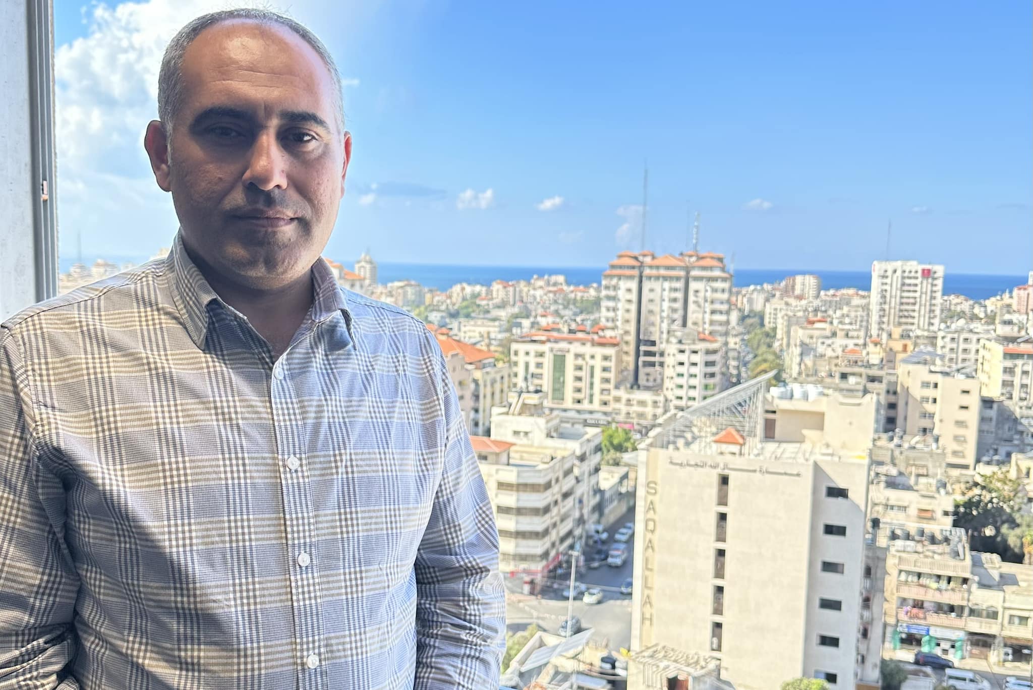 A middle aged man stands in front of a window, with a city skyline in the background. 