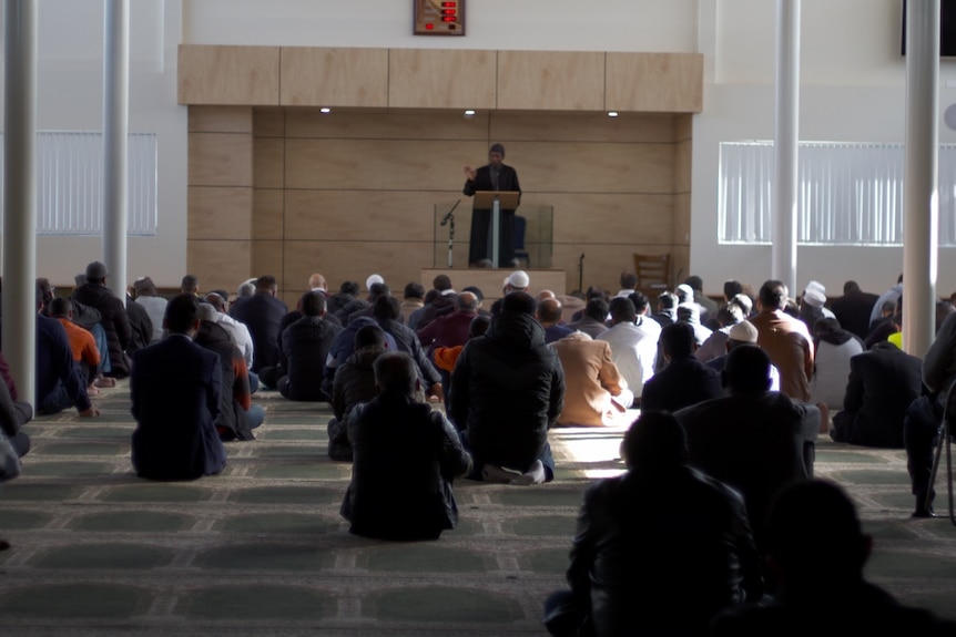 Worshippers sit inside a mosque and listen to an imam.