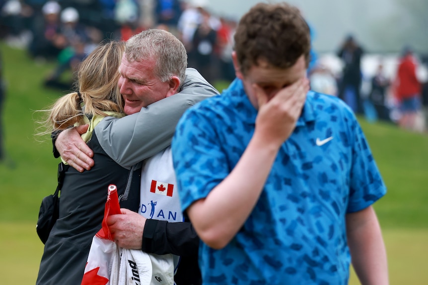 Golfer Robert MacIntyre, blurry in the foreground, cries as he walks away from his dad and caddy hugging his girlfriend.