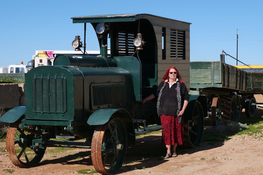A lady in a black jumper and red skirt stands in front of a dark green truck with two trailers made of wood.