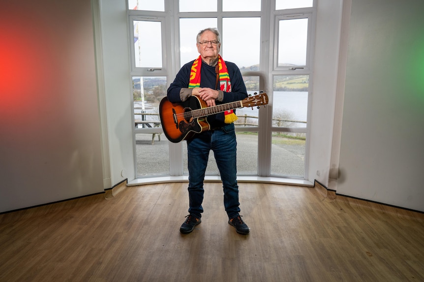 A man stands in front of a window holding a guitar.