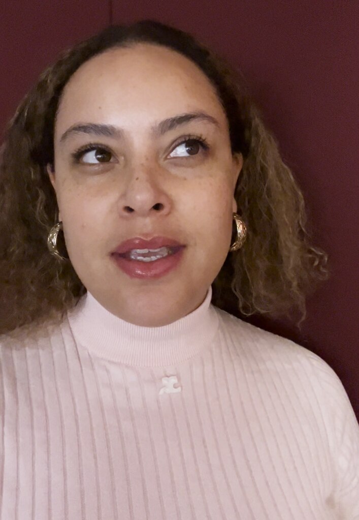 Tanya wears a light pink sweatshirt and gold hooped earrings. She's standing in front of a dark wall.