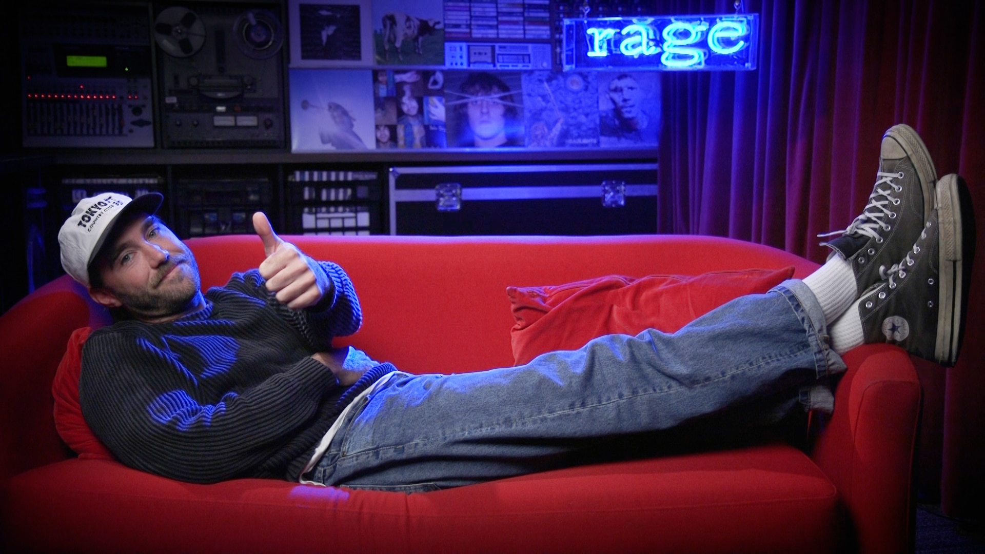 matt corby is doing a thumbs up while laying on a red couch. He sits in front of a neon blue sign that reads rage