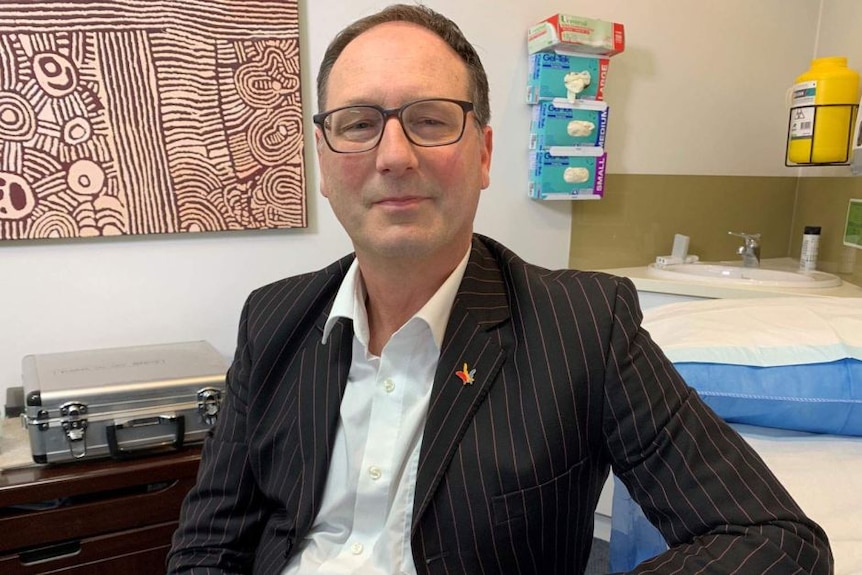 A bespectacled man in a dark blazer sitting in an office.