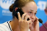 Ash Barty, sitting at a press conference, speaks on a mobile phone. She has her finger in her other ear.