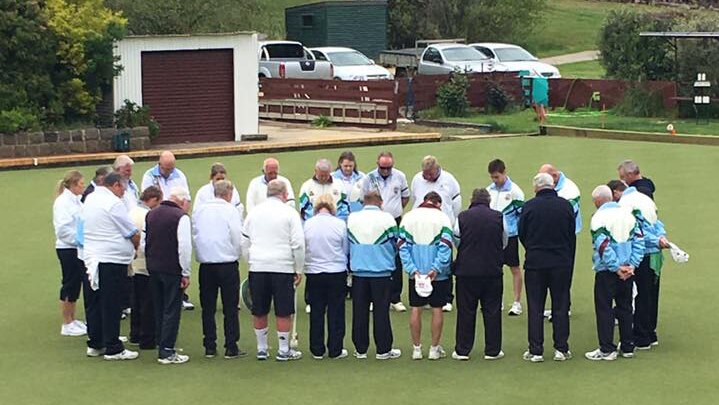 The Mt Xavier Bowling Club stand in a circle on the bowling green, heads bowed.