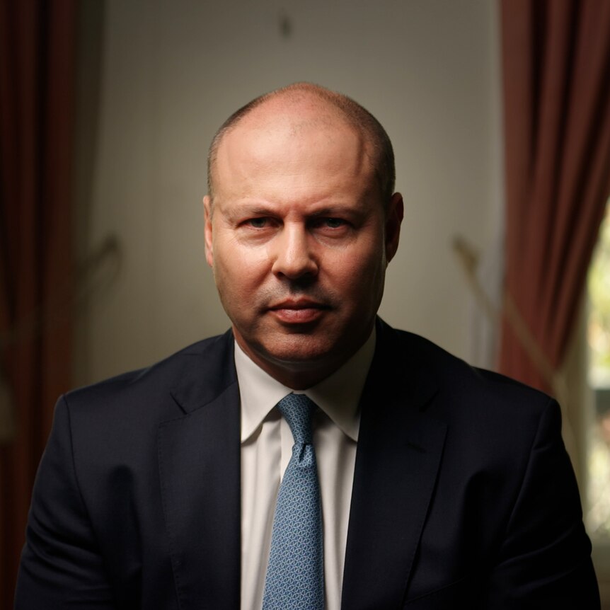 Dressed in a suit and tie, Josh Frydenberg sits in a room, looking into camera with a serious facial expression.