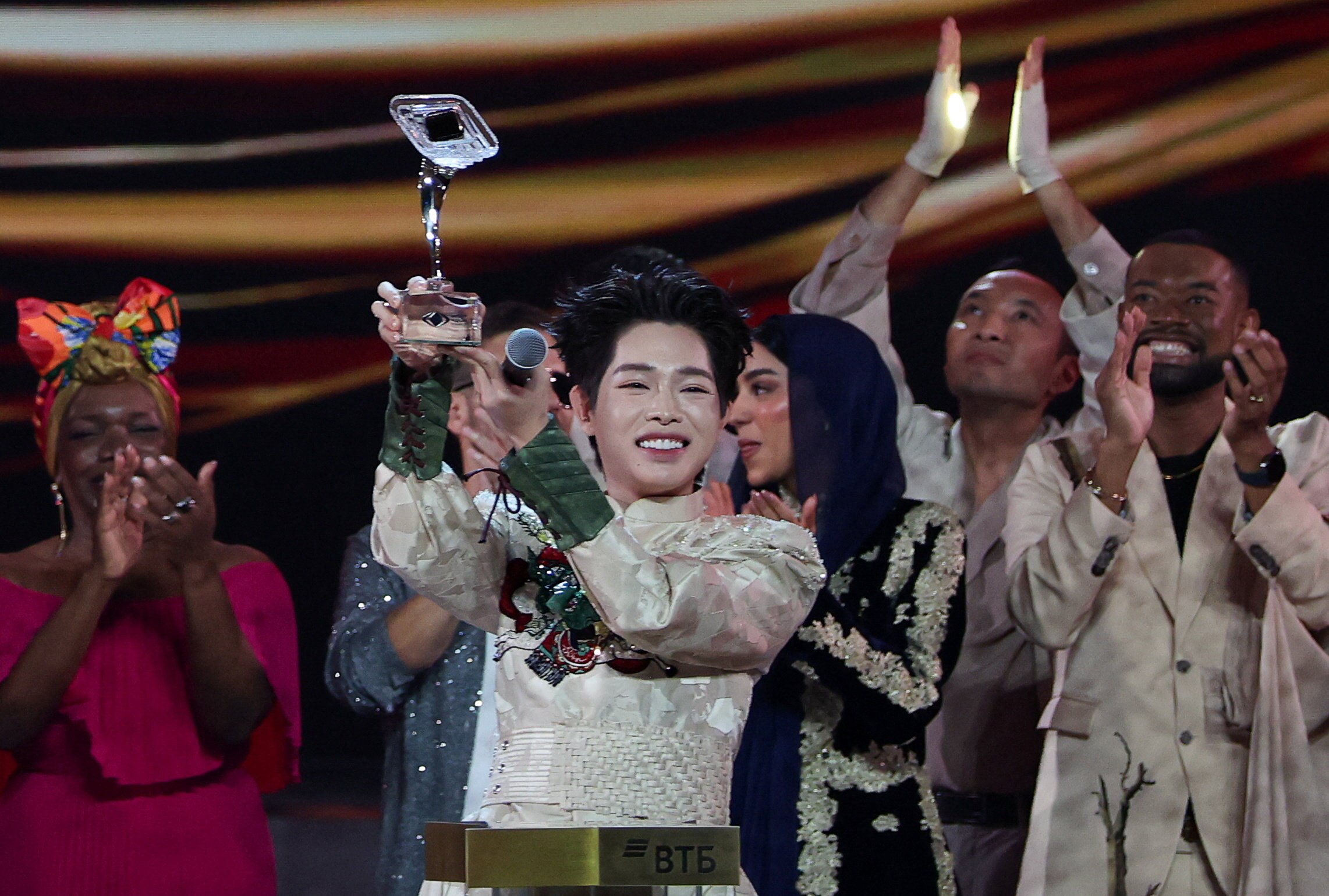 A man in a cream silk outfit holds up a trophy while surrounded by cheering people