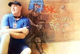 Zak Grieve, wearing a prison ID tag, baseball cap and sunglasses around his neck, sits in front of a wall with art on it.