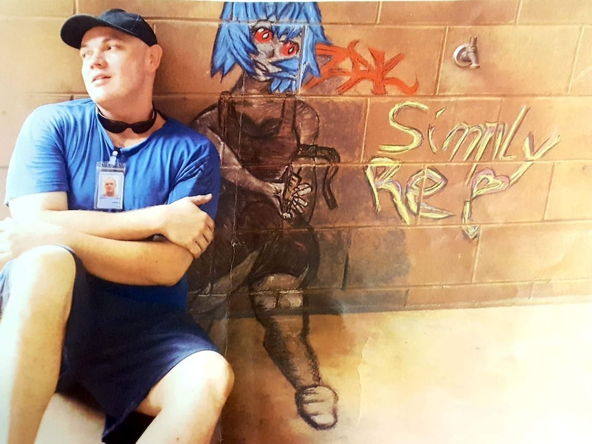 Zak Grieve, wearing a prison ID tag, baseball cap and sunglasses around his neck, sits in front of a wall with art on it.