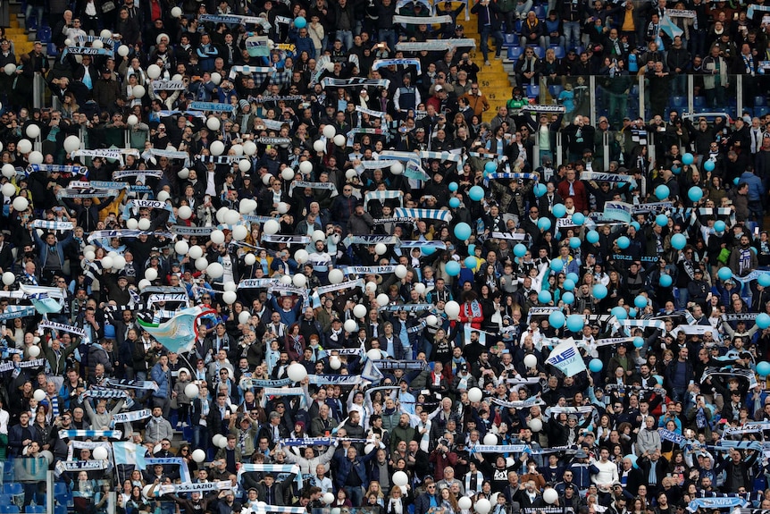 A big crowd of Italian football fans in a grandstand hold blue and white scarves and balloons