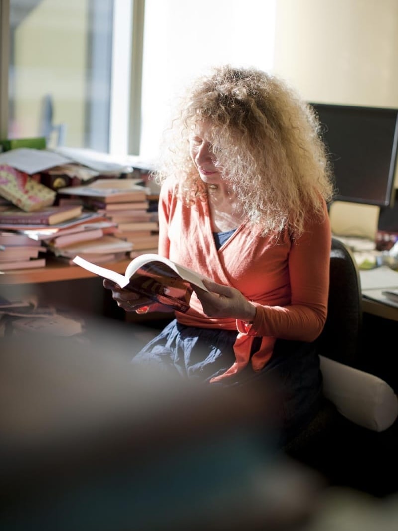 Kohn at desk reading book with pile of books in background.