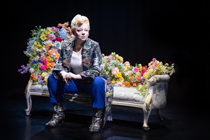 Sarah Snook, dressed in an ornate coat and wig, sits on a chair adorned by flowers, looking off to the side.