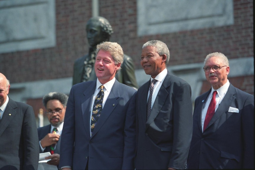 Bill Clinton and Nelson Mandela, dressed in grey suits, stand side by side in front of a statue