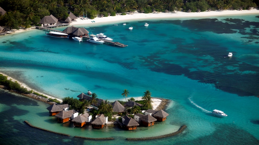 An aerial shot of a tropical blue lagoon, with beachfront bungalows, resorts and yachts.