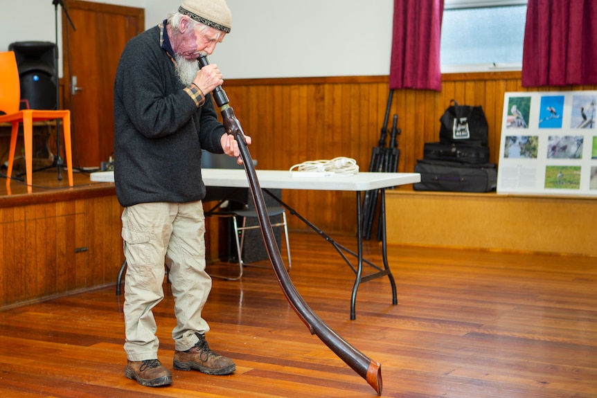 An aged man playing a leather didgeridoo in a small wooden-floored hall.